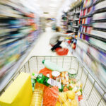 View over filled shopping cart racing down a supermarket aisle towards an out-of-focus woman shopper with extreme motion blur transforming into rainbow streaks along the shelves. NOTE: The product labels in the cart are dummies made by the photographer.