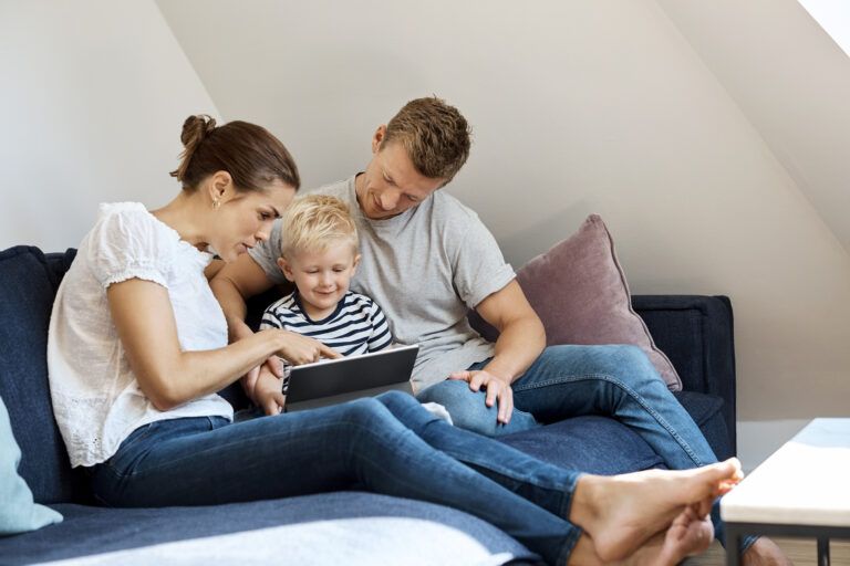 Loving parents with little son using digital tablet on sofa. Happy family of three spending leisure time at home. They are wearing casuals.