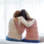 Rear back view of a mother and daughter embrace sitting on bed at home, older sister consoling younger teen, girl suffers from unrequited love share secrets trustworthy person relative people concept