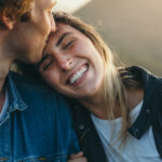 Close-up of romantic young boyfriend kissing smiling beautiful girlfriend on forehead while enjoying weekend at sunset